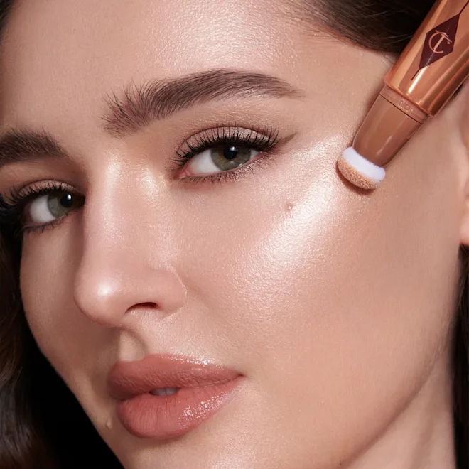 Close-up of a woman's face as foundation is applied with a makeup sponge to her cheek, showing dewy, glowing skin.