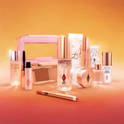 251001_Summer_Sale_26_sl_wk_202605_TravelMinisBagCreativeStillProductsinfrontofBag_R4_1000-x-1000jpg - shopcosmoprof.com Assorted pink-and-gold makeup products arranged on a peach background, including a compact, bottles, and brushes.