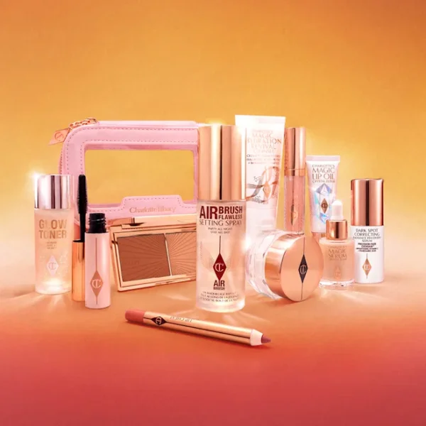 Assorted pink-and-gold makeup products arranged on a peach background, including a compact, bottles, and brushes.