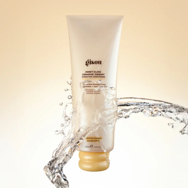 Gisou Honey Gloss conditioner tube with a yellow cap, surrounded by a splash of water against a pale background.