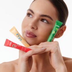 Woman holding three Lipss lip balm tubes (red, yellow, green) near her face against a white background.