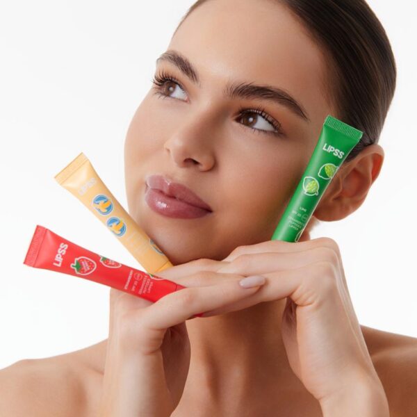 Woman holding three Lipss lip balm tubes (red, yellow, green) near her face against a white background.