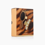 Balmain Paris hair styling set box with a black round brush and gold-toned styling tool against a sandy gold background (retail packaging)