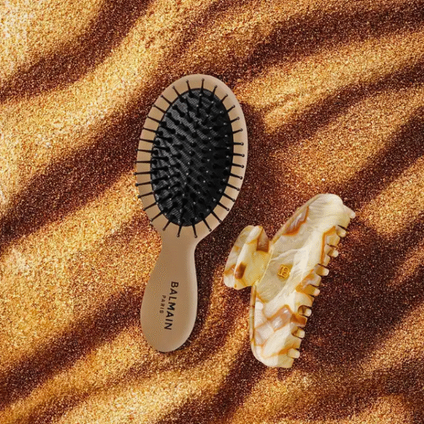 Two hair brushes resting on a sandy surface: a Balmain Paris paddle brush with black bristles and a beige tortoiseshell comb beside it on the sand.