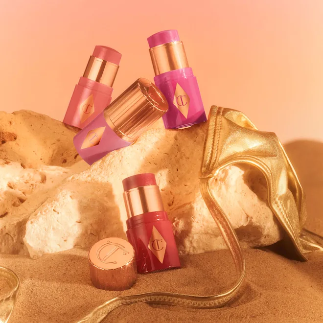 Assorted pink and gold lipsticks arranged on a rocky sand backdrop with a gold purse in the scene.