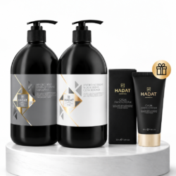 Hadat hair and hand care set displayed on a white marble pedestal: two black pump bottles (Shampoo and Conditioner) and two Hadat Calm Hand Cream tubes with packaging and a gift badge nearby.