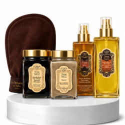 Luxurious spa gift set on a white marble pedestal: two gold spray bottles, two cream jars with gold lids, and a brown exfoliating mitt in the background.