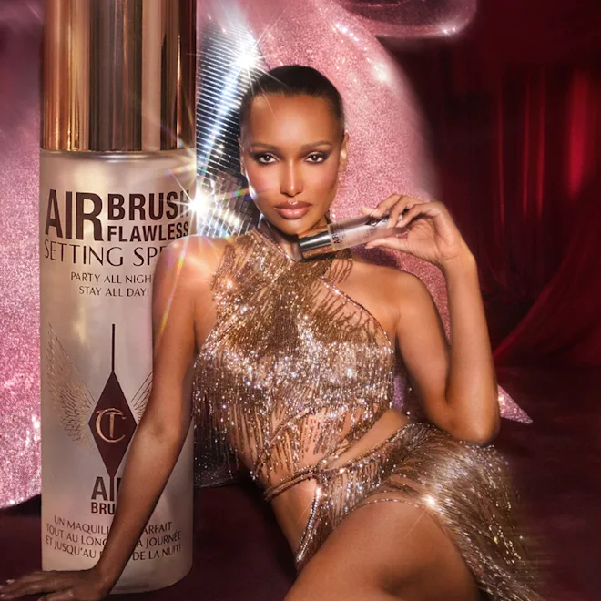 Model in a sparkling gold outfit posing with a large can of Airbrush Flawless Setting Spray in a pink, glittery backdrop.