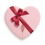 Heart-shaped pink quilted gift box tied with a red ribbon bow and tag showing a brand logo