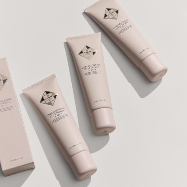 Pink hair primer tubes arranged on a pale background, product label reads 'Hair Discipline Style Primer 5‑In‑1' (HADAT). Scientific/cosmetic styling product promo image.